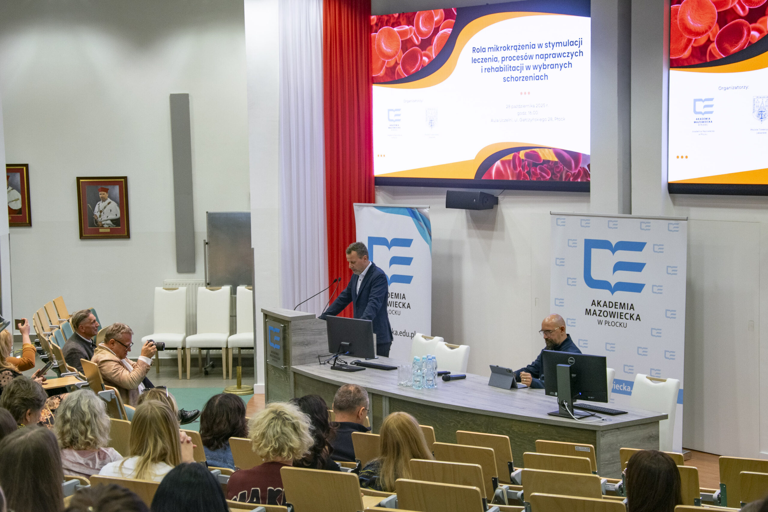 Mężczyzna stoi przy mównicy , drugi siedzi przy biurku z tabletem, za nimi ekrany z prezentacją