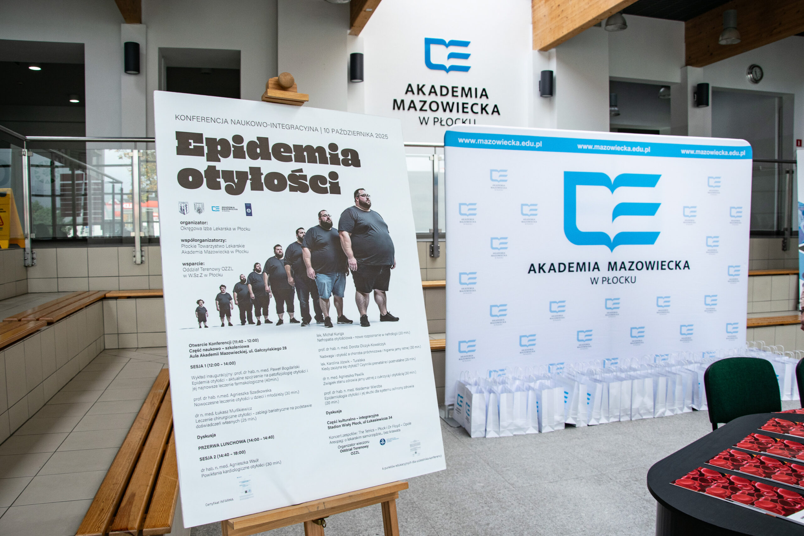 widok na plakat promujący konferencję naukową "Epidemia otyłości", ustawiony na sztaludze, w tle ścianka z logotypem Akademii Mazowieckiej w Płocku