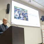 Jarosław Wanecki stojący przy mównicy - prelegent a za nim pokaz slajdów i rollapy